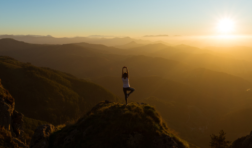 A young woman does a tree yoga pose while standing on a small mountain overlooking rolling foothills as the sun sets